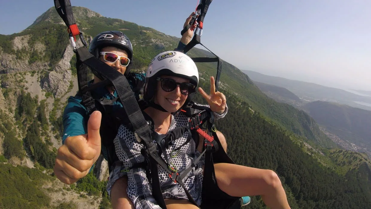 Passenger seated in a tandem paragliding harness above the Kotor landscape.